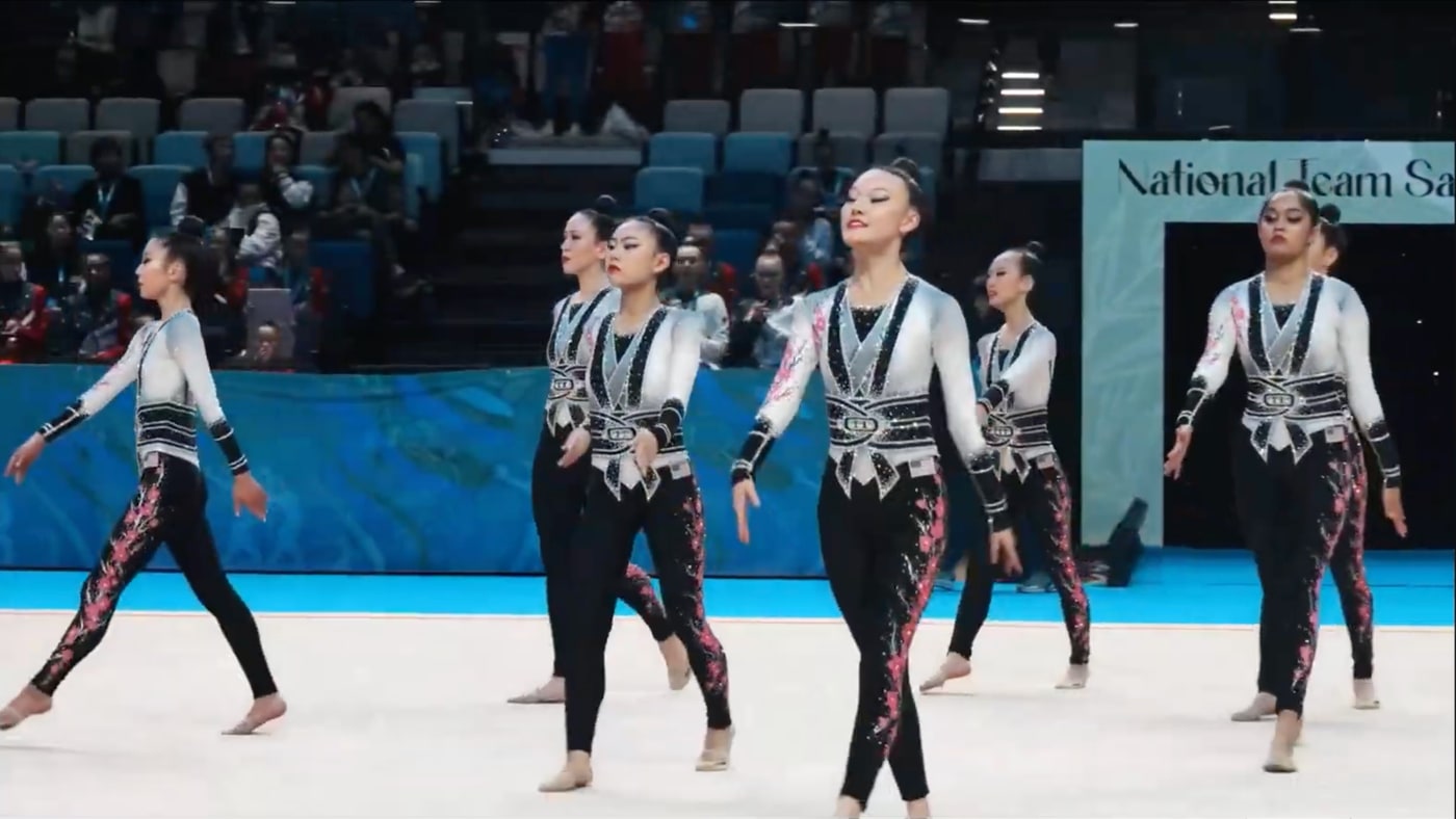 Seven gymnasts performing together in matching uniforms on a competition floor.